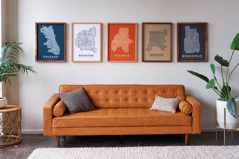 May include: Five framed city maps of Chicago, Houston, Atlanta, Denver, and Minneapolis hang on a white wall above a brown leather sofa with two throw pillows. A potted plant is on the right side of the image and a small wicker table is on the left.