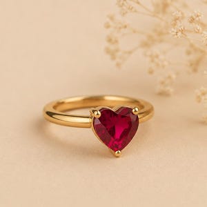 May include: A gold-toned ring featuring a single heart-shaped red gemstone set in a prong setting. The band is smooth and polished, showcasing a minimalist design that highlights the vibrant crimson color of the central heart-shaped stone.