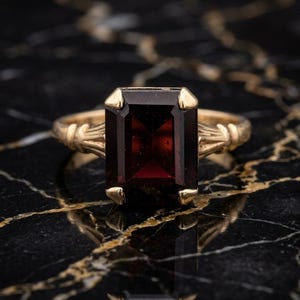 Könnte beinhalten: Ein Goldring mit einem großen, rechteckigen, im Smaragdschliff geschliffenen, dunkelroten Granat-Edelstein, der von vier Krappen gehalten wird. Die Ringschiene zeigt auf beiden Seiten ein dekoratives, strukturiertes Metalldesign.