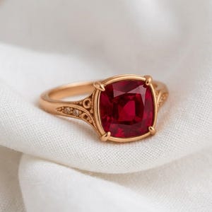 Peut inclure: Une bague en or ornée d'une grande pierre précieuse rouge foncé de taille coussin, fixée par quatre griffes. L'anneau présente des détails de volutes gravées complexes sur les côtés, menant au sertissage de la pierre centrale.