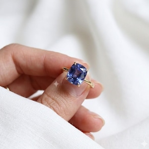 Può includere: Un anello in oro con una grande pietra preziosa zaffiro blu taglio a cuscino, montata su una struttura a quattro griffe. La fascia è sottile e lucida, mettendo in risalto il colore blu intenso e vibrante della pietra sfaccettata.