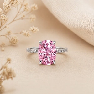 May include: A silver ring featuring a large, oval-cut pink gemstone. The band is embellished with smaller, clear gemstones. The ring is displayed against a neutral backdrop, with soft lighting.