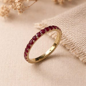 May include: A gold ring featuring a continuous row of small, round, red gemstones. The ring is displayed on a neutral-toned surface, with a sprig of dried flowers in the background. The ring's design is simple and elegant.