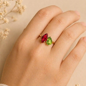 May include: A gold ring featuring two teardrop-shaped gemstones, one red and one green. The ring is displayed on a hand, with a neutral background and small floral accents.