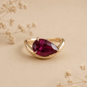 Puede incluir: Un anillo de oro con una gran piedra preciosa roja oscura en forma de pera. El anillo tiene una banda lisa y redondeada, y la piedra preciosa está sujeta por pequeñas garras. El anillo se muestra sobre un fondo suave y neutro, con delicados acentos florales.