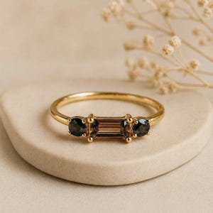 May include: A gold ring with three gemstones. Two dark, round stones sit either side of a rectangular, brown stone. The ring is displayed on a neutral surface, with a blurred floral element in the background.