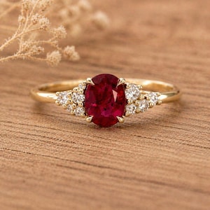 Può includere: Un anello d'oro con un grande rubino ovale come pietra centrale, affiancato da pietre preziose rotonde e trasparenti più piccole. L'anello è posto su una superficie di legno, mettendo in risalto il design e il colore del gioiello.