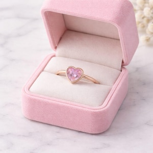 Puede incluir: Un anillo de tono dorado con una gema rosa en forma de corazón engastada en un bisel, presentado dentro de un joyero cuadrado de terciopelo rosa claro con un forro interior de color crema diseñado para mantener el anillo en su lugar.