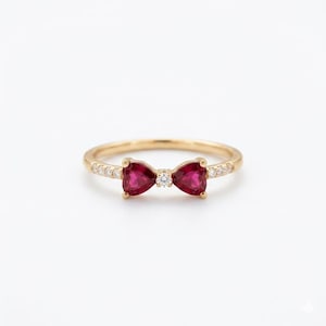 May include: A gold ring featuring a bow design. The bow is made of two heart-shaped, deep red gemstones with a small, round diamond in the center. Small diamonds are set along the band on either side of the bow.