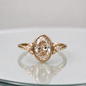 Puede incluir: Un anillo de oro con una gran piedra central de corte ovalado en color champán engastada en un bisel liso. La banda está acentuada a cada lado por un grupo de tres pequeñas piedras redondas y transparentes, creando un diseño equilibrado.
