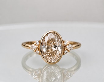 May include: A gold ring featuring a large, oval-cut champagne-colored center stone set in a smooth bezel. The band is accented on each side by a cluster of three small, round, clear stones, creating a balanced and symmetrical design.