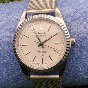May include: Silver HMT Kohinoor wristwatch with a mesh strap. The watch face is white with black hands and hour markers, and the text "PARA SHOCK 17 JEWELS" is visible. The watch is set against a blue background.