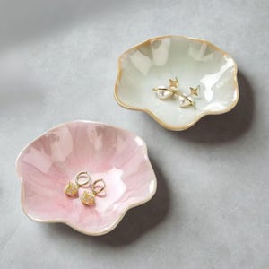 以下が含まれることがあります： 2つの装飾的な花びら型の小物入れ。1つは淡いピンク色、もう1つはオフホワイト色です。どちらも金色の縁取りがあります。ピンク色の皿には、正方形のテクスチャードチャームが付いたゴールドフープイヤリングが入っています。オフホワイトの皿には、パールと星のアクセントが付いたゴールドイヤリングが入っています。