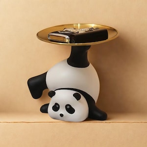 Puede incluir: Una bandeja decorativa con forma de panda con una parte superior circular dorada. El panda es blanco y negro, con un pequeño cachorro de panda en la base. La bandeja contiene un juego de llaves y un objeto negro.