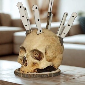 May include: A skull-shaped knife block, holding several knives. The skull is beige with a weathered look, and the knives have white handles with black dots. The base is a wooden slice. A unique kitchen accessory.