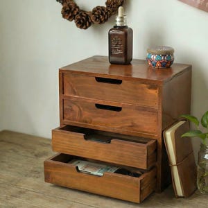 以下が含まれることがあります： 濃い茶色の仕上げの、3段の木製収納ボックス。 ボックスの上には、スポイト付きの茶色のボトルと小さな装飾ジャーがあります。 1つの引き出しが開いており、中身が見えます。 革張りのノートがボックスの横に置かれています。