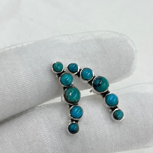 May include: A pair of silver earrings featuring a curved design set with multiple round turquoise stones. Each earring displays a graduated arrangement of blue-green cabochons held in place by silver settings, creating a distinct arc shape.