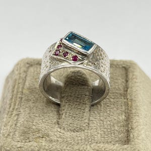 May include: A silver ring with a wide, hammered band featuring a rectangular blue gemstone set horizontally. Next to the blue stone, a triangular silver accent is embedded with four small, round, deep pinkish-red gemstones arranged in a row.