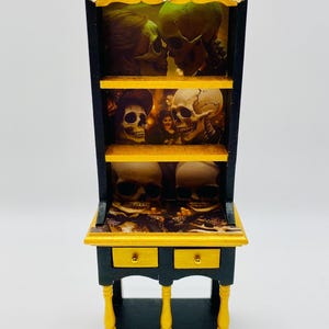 May include: Miniature black and gold cabinet with shelves and drawers. The cabinet is decorated with images of skulls. The top and drawer fronts are gold, and the legs are gold with black accents. Decorative gold accents are on the top.