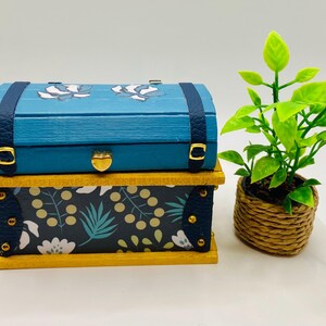 May include: A miniature blue wooden chest with a floral design on the lid and a patterned base. The chest has gold-colored hardware and dark blue straps. A small potted plant with green leaves sits next to the chest.