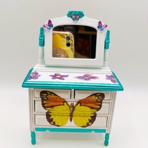 May include: A white miniature dresser with a mirror, decorated with butterfly and floral designs. The dresser has a large yellow and orange butterfly on the drawer fronts, with teal accents and purple flower details.