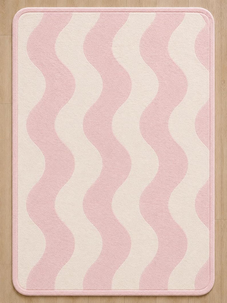 May include: A rectangular area rug featuring a repeating vertical wavy pattern in soft pink and cream colors. The rug has rounded corners and a solid pink border that frames the entire design, creating a clean and consistent aesthetic throughout.
