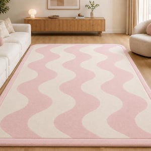 May include: A rectangular area rug featuring a repeating wavy pattern in soft pink and cream colors. The rug has rounded corners and a solid pink border that frames the entire design, creating a clean and defined edge for the floor covering.