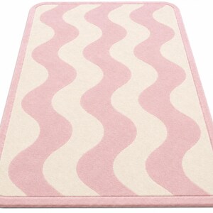 May include: A rectangular area rug featuring a repeating vertical wavy pattern in soft pink and cream colors. The rug has a solid pink border that frames the entire design, with rounded corners and a consistent texture across the surface.