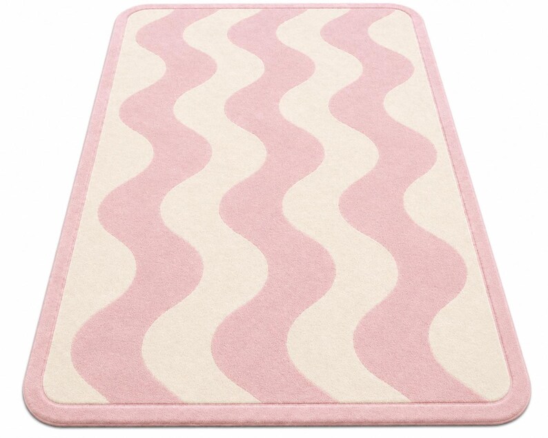 May include: A rectangular area rug featuring a repeating vertical wavy pattern in soft pink and cream colors. The rug has a solid pink border that frames the entire design, with rounded corners and a consistent texture across the surface.