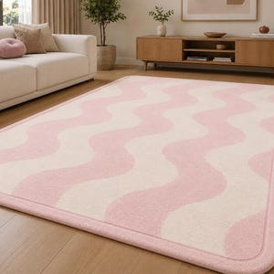 May include: A rectangular area rug featuring a repeating pattern of soft pink and cream-colored wavy vertical stripes. The rug has rounded corners and a solid pink border that frames the entire design, lying flat on a light-colored wooden floor.