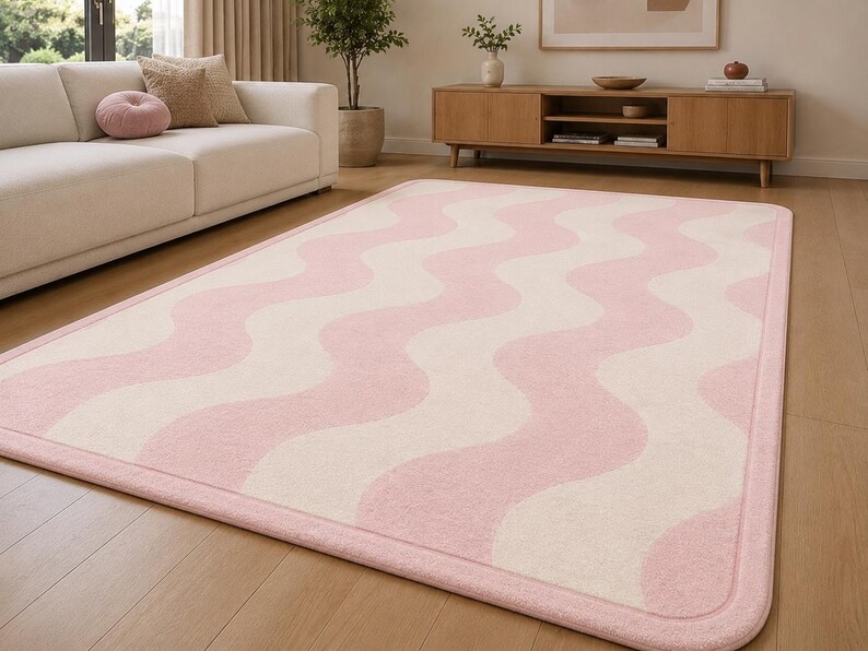 May include: A rectangular area rug featuring a repeating pattern of soft pink and cream-colored wavy vertical stripes. The rug has rounded corners and a solid pink border that frames the entire design, lying flat on a light-colored wooden floor.