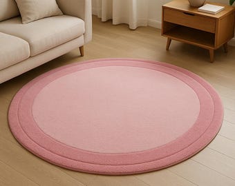 May include: A round, pink rug with a layered design. The rug features a light pink center, surrounded by a medium pink band, and a darker pink border. The rug is placed on a light-colored wooden floor.