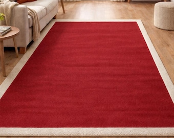Pode incluir: Um tapete retangular vermelho com uma borda creme é colocado sobre um piso de madeira. O tapete é o ponto focal de uma sala de estar, com um sofá, mesa lateral e outros móveis visíveis ao fundo, criando um ambiente acolhedor.