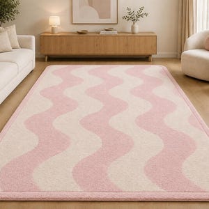 May include: A rectangular area rug featuring a repeating wavy pattern in soft pink and cream colors. The rug has rounded corners and a solid pink border that frames the entire design, creating a clean and cohesive look for the floor covering.