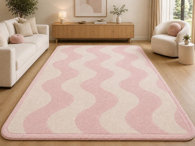 May include: A rectangular area rug featuring a repeating wavy pattern in soft pink and cream colors. The rug has rounded corners and a solid pink border that frames the entire design, creating a clean and cohesive look for the floor covering.
