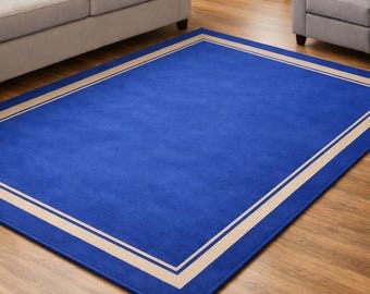 Pode incluir: Um tapete azul retangular com uma borda bege. O tapete é colocado em um piso de madeira e é cercado por móveis. O design do tapete é simples e moderno, adequado para salas de estar ou quartos.
