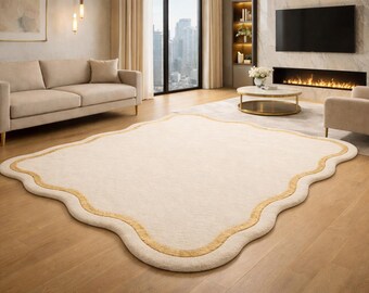 May include: A cream-colored, scalloped-edge area rug with a gold border. The rug is placed on a hardwood floor in a living room setting, with a sofa, armchair, and coffee table visible.