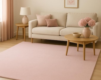 Op de afbeelding: Een lichtroze rechthoekig vloerkleed bedekt de vloer in een woonkamer. Een beige bank met roze kussens, een houten salontafel en een bijzettafel met een lamp zijn ook aanwezig. Een vaas met roze bloemen staat op de salontafel.