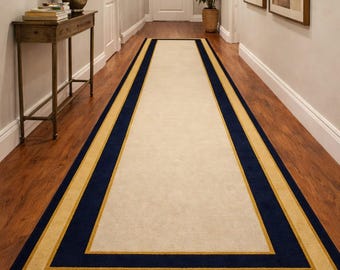 Pode incluir: Um tapete retangular para corredor com um centro creme, ladeado por uma faixa azul marinho e uma faixa dourada. O tapete é colocado sobre um piso de madeira em um corredor. O tapete adiciona um elemento decorativo ao espaço.