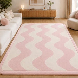 May include: A rectangular area rug featuring a repeating wavy pattern in soft pink and cream colors. The rug has rounded corners and a solid pink border that frames the entire design, creating a clean and consistent look for floor coverage.