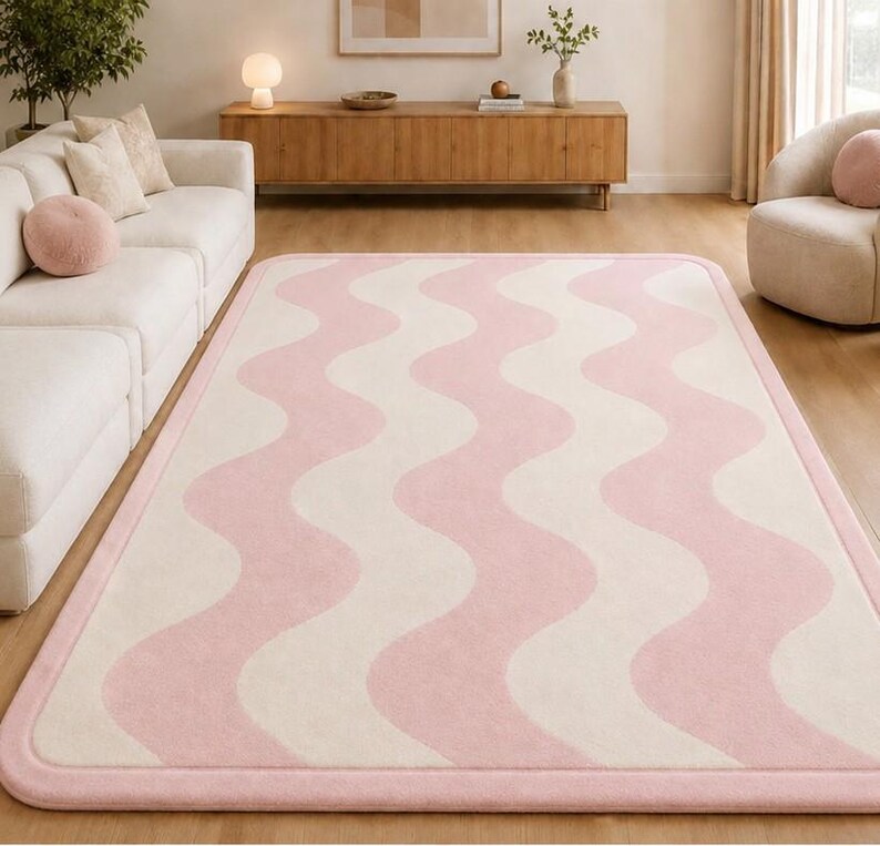 May include: A rectangular area rug featuring a repeating wavy pattern in soft pink and cream colors. The rug has rounded corners and a solid pink border that frames the entire design, creating a clean and consistent look for floor coverage.