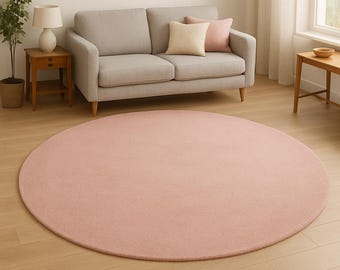 Pode incluir: Um tapete grande e redondo, de cor rosa pálido, domina o primeiro plano. O tapete é feito de um material macio e assenta num chão de madeira clara. Ao fundo, um sofá cinzento com almofadas e uma mesa lateral com uma lâmpada.