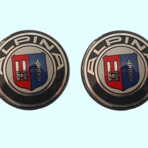May include: Two circular Alpina emblems. Each emblem features a silver border with the word "ALPINA" in capital letters. The center design is a shield divided into red and blue sections, with a detailed emblem in each.