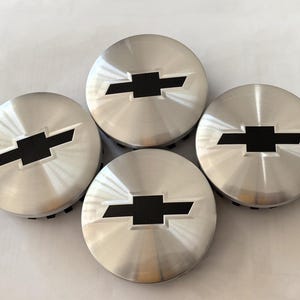 May include: Four silver-colored hubcaps with a black Chevrolet bow tie logo in the center. The hubcaps have a brushed metal finish and are arranged in a circular pattern against a white background.