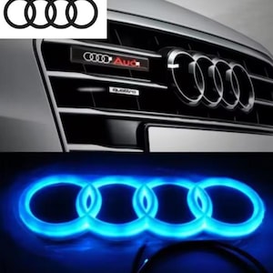 May include: An image showcasing the Audi logo in multiple forms. The top shows the classic interlocking rings on a car grille, with the Audi and Quattro emblems. Below, a glowing blue version of the logo is displayed.
