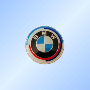 May include: A circular BMW badge with a silver edge. The badge displays the BMW logo, a blue and white quadrant within a black ring, encircled by red and blue rings, and the letters "B", "M", and "W".