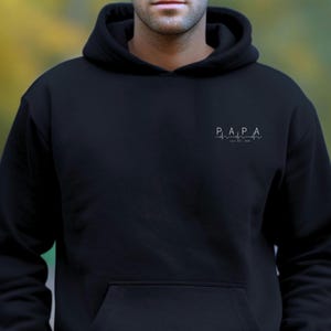 Puede incluir: Sudadera con capucha negra con bolsillo delantero. En el pecho aparece la palabra "PAPA" sobre un diseño de línea de latido cardíaco, con "Est. 2023" debajo. La sudadera es de color negro liso.
