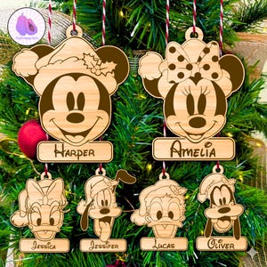 May include: Wooden Christmas ornaments featuring cartoon characters Mickey Mouse, Minnie Mouse, Donald Duck, Daisy Duck, Goofy, and Pluto. Each ornament has a name on a small wooden plaque. The ornaments are hanging on a Christmas tree.