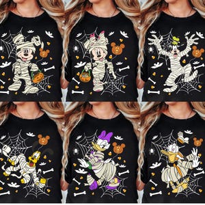 Pode incluir: Uma t-shirt preta com uma estampa gráfica de personagens da Disney enrolados em bandagens brancas como múmias. O design inclui teias de aranha, morcegos, ossos, doces de milho e ícones em forma de abóbora espalhados pelo tecido.