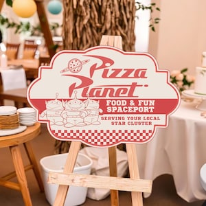 May include: A decorative sign on a wooden easel featuring the Pizza Planet logo in red and cream. The sign displays three green aliens, the text Pizza Planet Food & Fun Spaceport Serving Your Local Star Cluster, and a red and white checkered border.